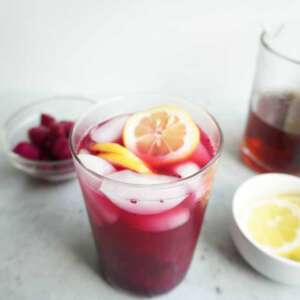A glass of low sugar dragon fruit lemonade with another bowl of lemon and dragon fruit pieces and a pitcher of tea in the background