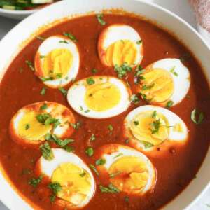 A white bowl filled with spicy red curry topped with sliced boiled eggs and garnished with chopped cilantro, with a side dish of fresh salad in the background.