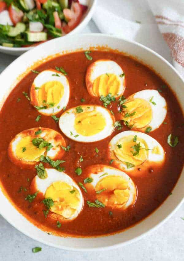 A white bowl filled with spicy red curry topped with sliced boiled eggs and garnished with chopped cilantro, with a side dish of fresh salad in the background.