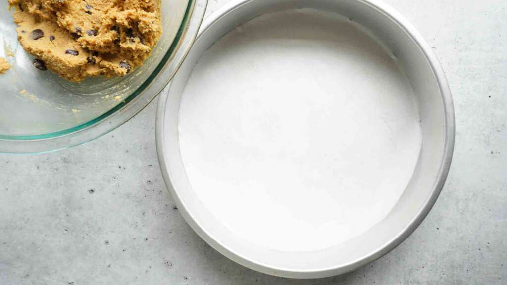 A bowl of cookie dough next to a parchment lined cake pan